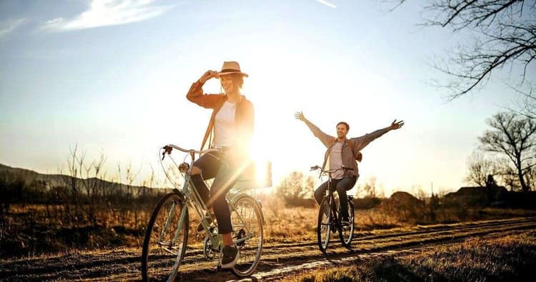 Άνδρας και γυναίκα κάνουν ποδήλατο σε χωμάτινο μονοπάτι στην ύπαιθρο κατά το ηλιοβασίλεμα· η γυναίκα μπροστά φορά καπέλο και χαμογελά, ενώ ο άνδρας πίσω απλώνει τα χέρια του χωρίς να κρατά το τιμόνι, με τον ήλιο να λάμπει έντονα στο φόντο.