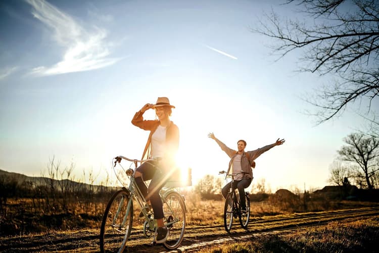 Άνδρας και γυναίκα κάνουν ποδήλατο σε χωμάτινο μονοπάτι στην ύπαιθρο κατά το ηλιοβασίλεμα· η γυναίκα μπροστά φορά καπέλο και χαμογελά, ενώ ο άνδρας πίσω απλώνει τα χέρια του χωρίς να κρατά το τιμόνι, με τον ήλιο να λάμπει έντονα στο φόντο.