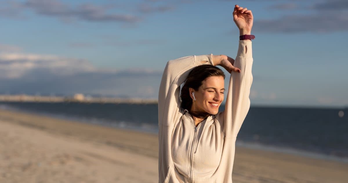 Μια νεαρή γυναίκα με κλειστά μάτια και ένα πλατύ χαμόγελο τεντώνει τα χέρια της πάνω από το κεφάλι της στην αμμουδιά. Φοράει μπεζ ζακέτα με κουκούλα, ασύρματα ακουστικά και ένα αθλητικό ρολόι, με φόντο την ήρεμη θάλασσα και τον γαλάζιο ουρανό.